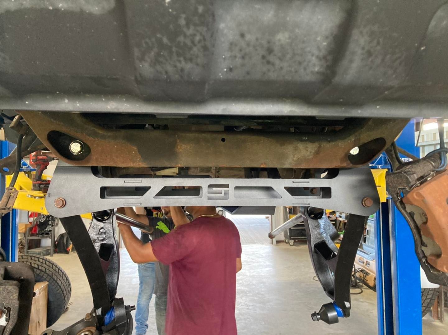 Two mechanics installing a grey suspension component on a vehicle lifted in a shop.