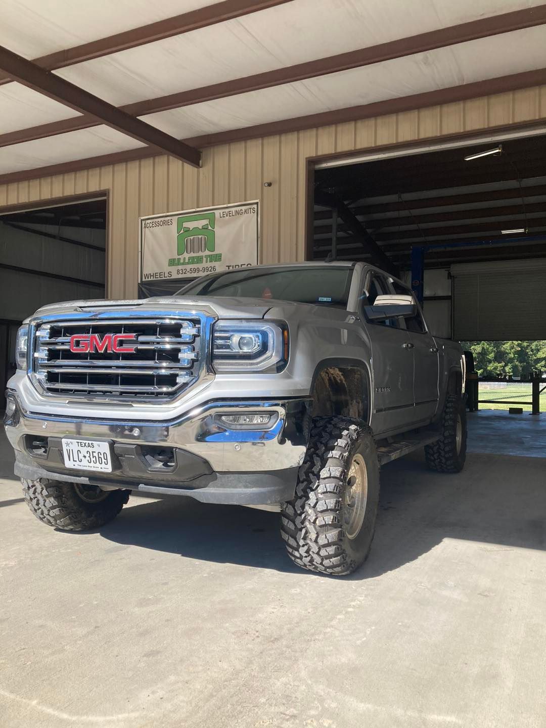 Silver GMC truck with large tires parked in a garage with open doors.