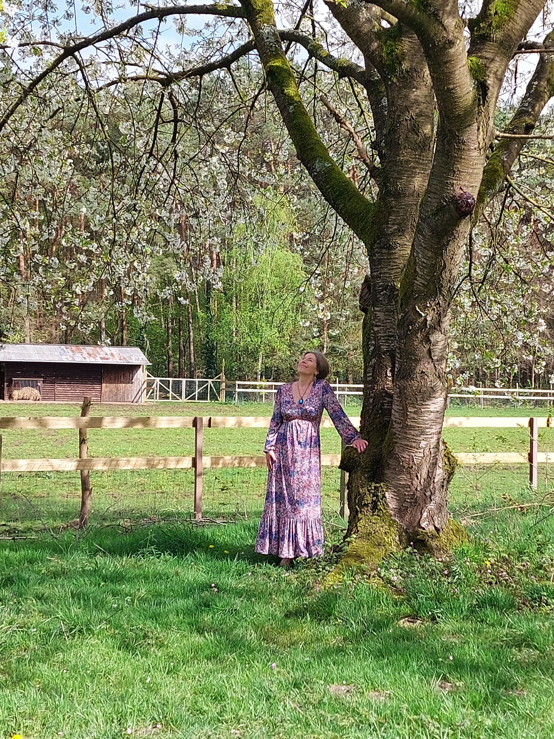 Een vrouw in een lange jurk staat naast een boom in een veld.