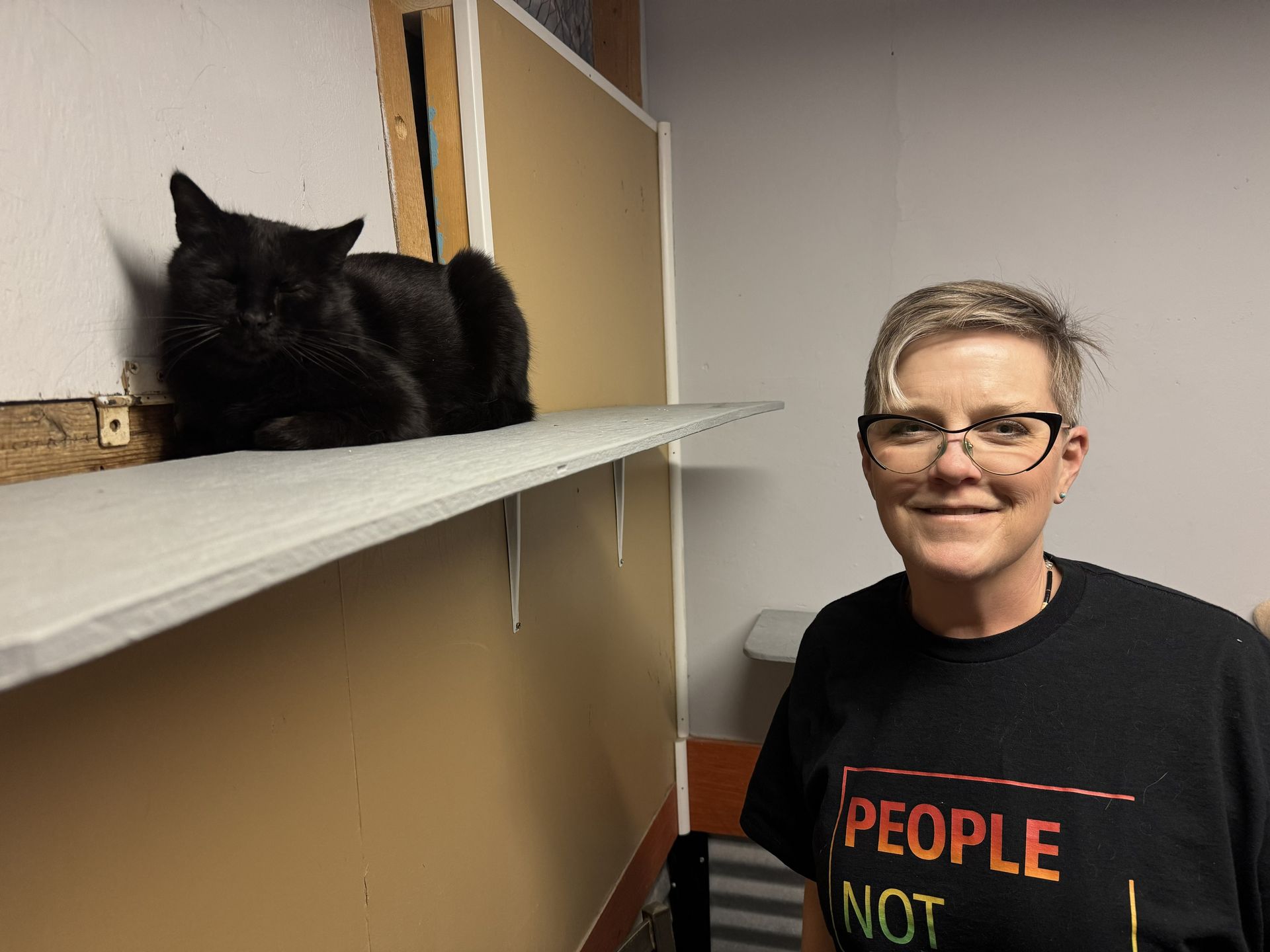 A person with short hair and glasses smiles while standing next to a black cat lounging on a wall-mounted shelf.