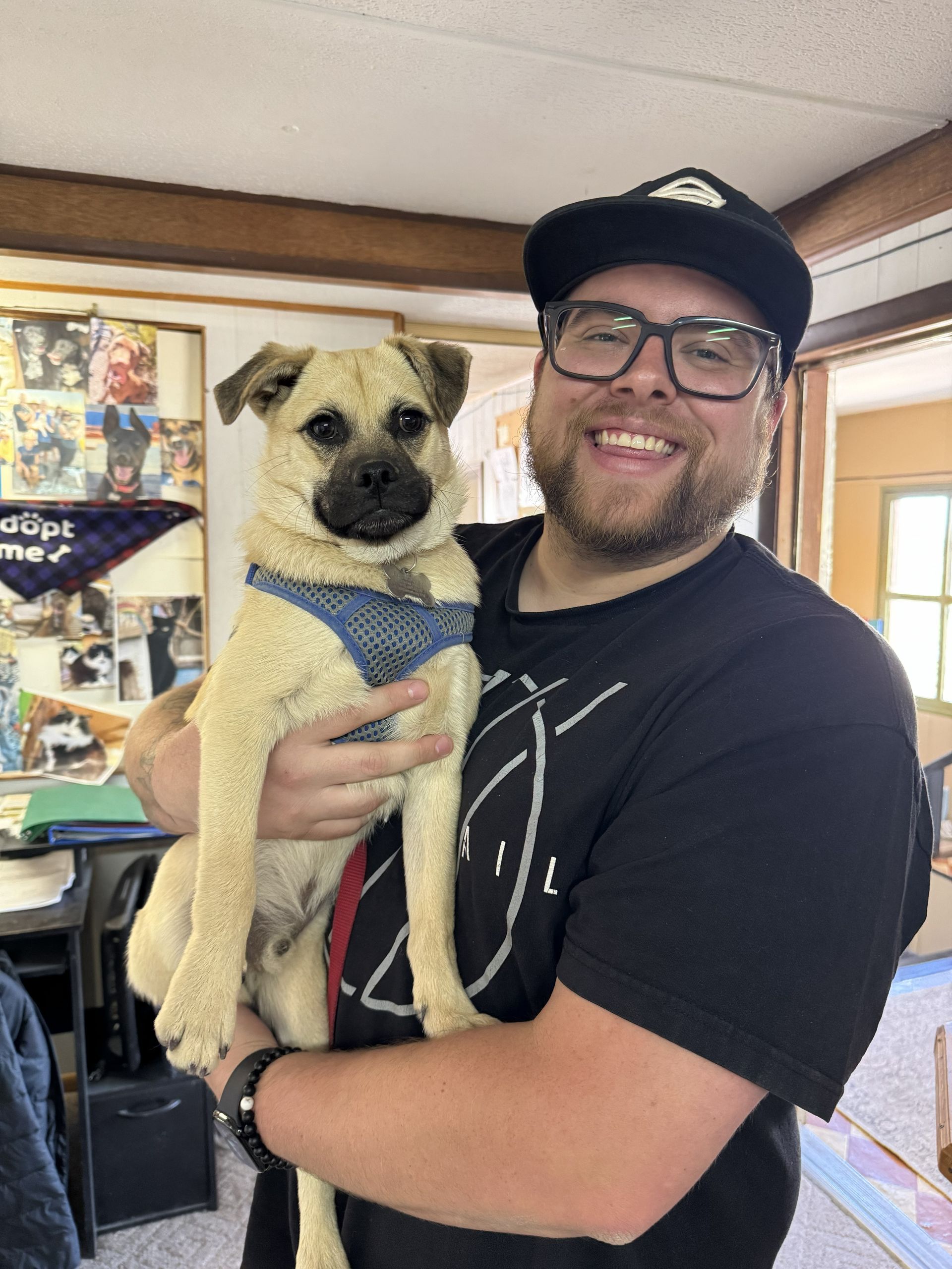 Man holding a small tan dog indoors, both facing the camera and wearing casual clothes.