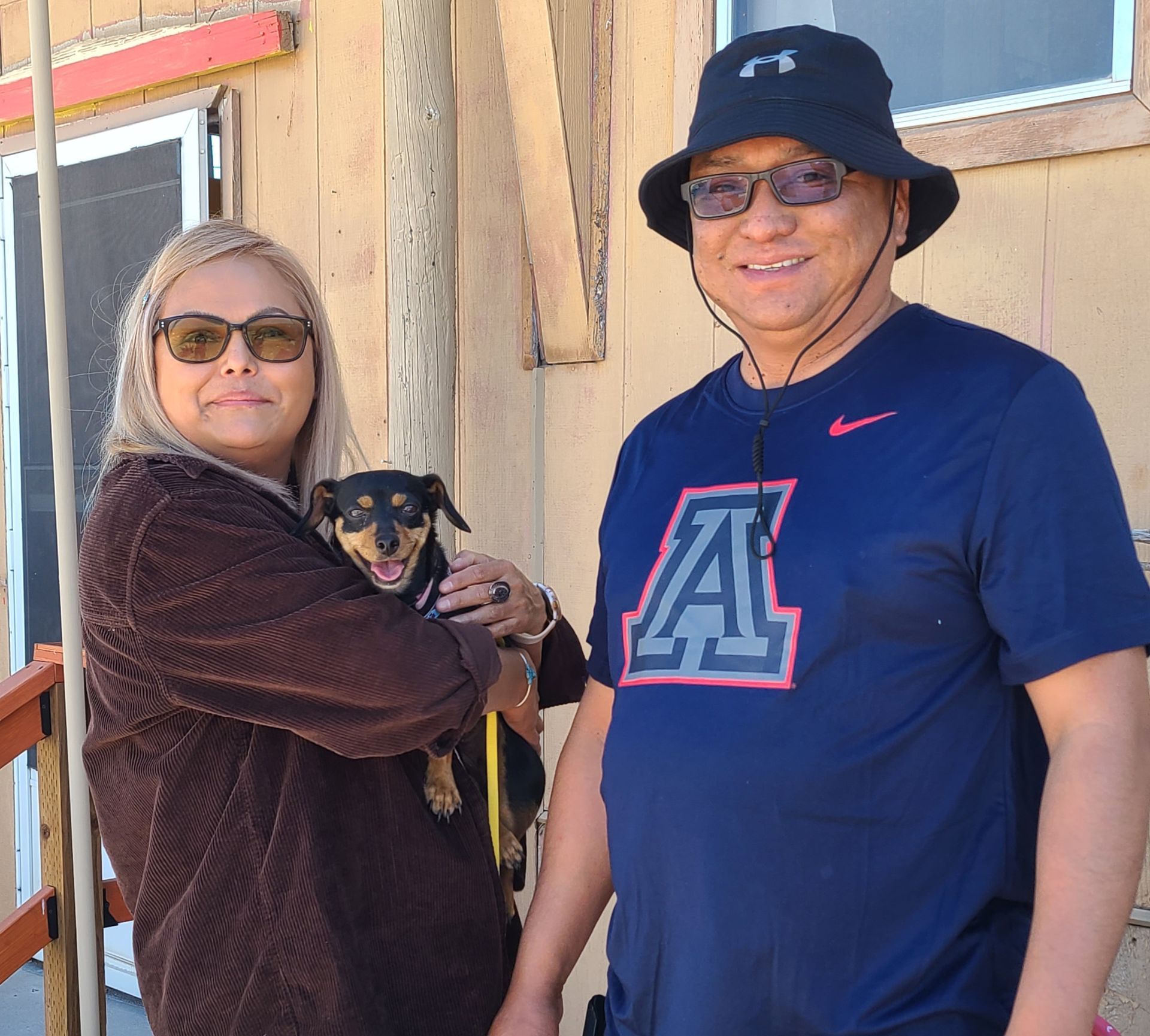 A smiling couple stands outdoors; the woman holds a small dog, and the man wears a dark blue t-shirt and bucket hat.