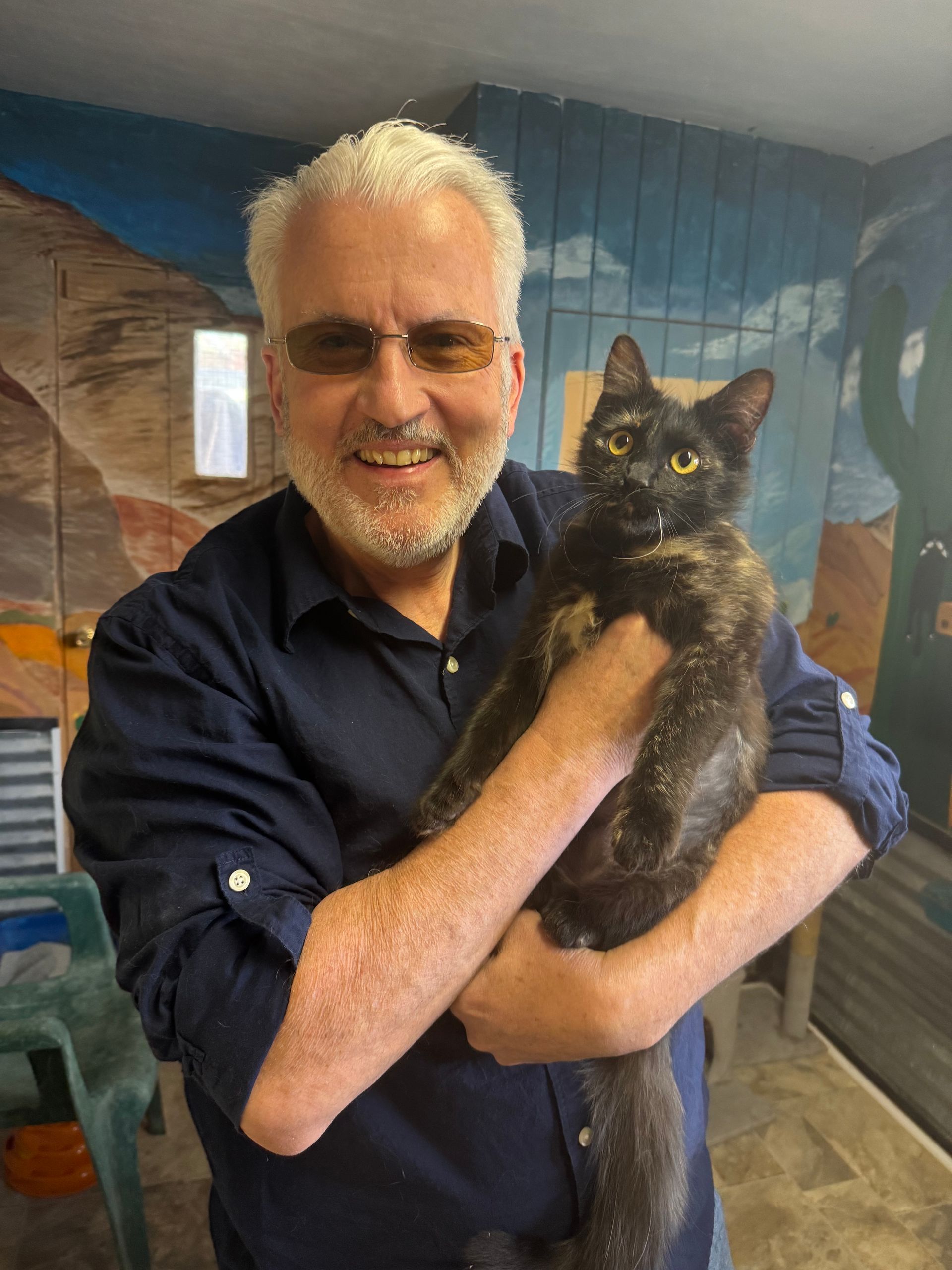 Man holding a black cat indoors, smiling in front of a mural backdrop