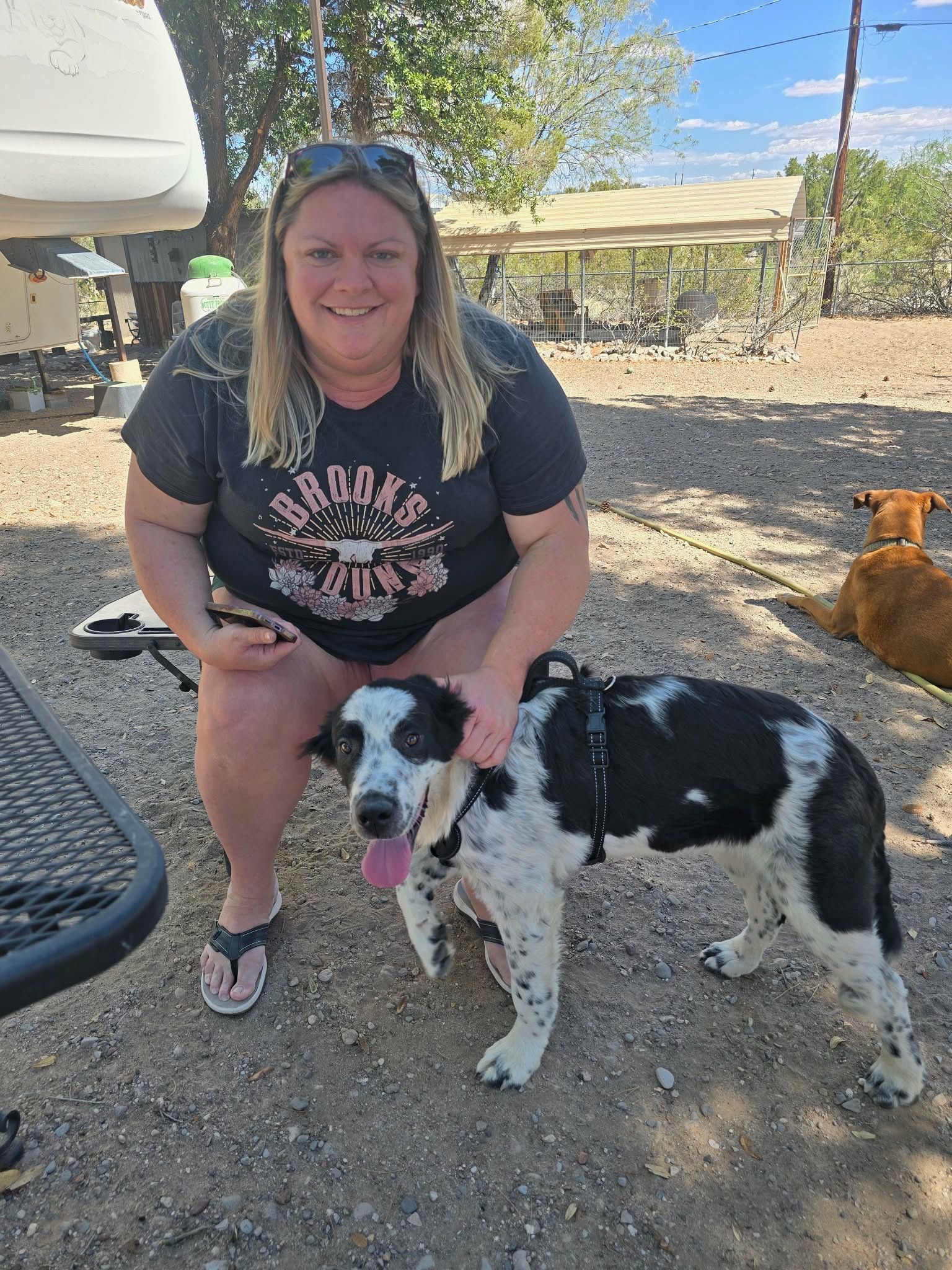 Woman kneeling beside a black-and-white dog outdoors, both facing the camera on a sunny dirt path.
