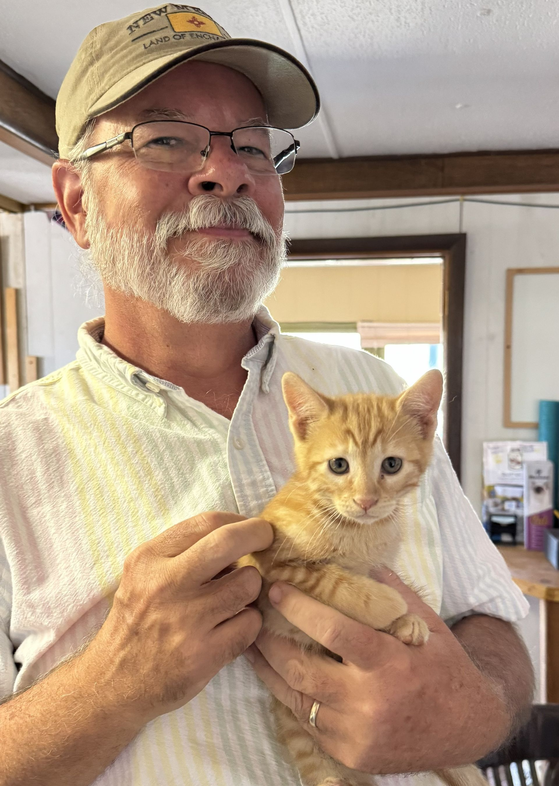 Man in a tan cap holding a ginger kitten indoors, both facing the camera