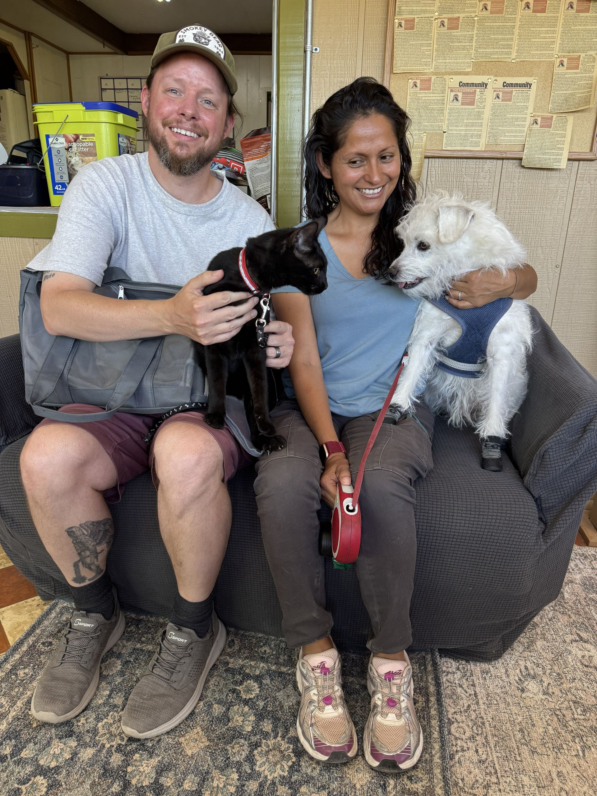 Two people on a couch holding a black dog and a white dog, smiling in a casual indoor setting.