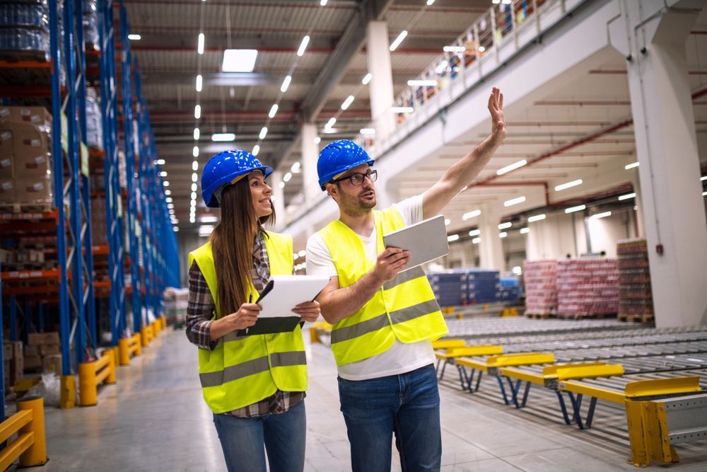 A man and a woman are standing in a warehouse looking at a tablet.