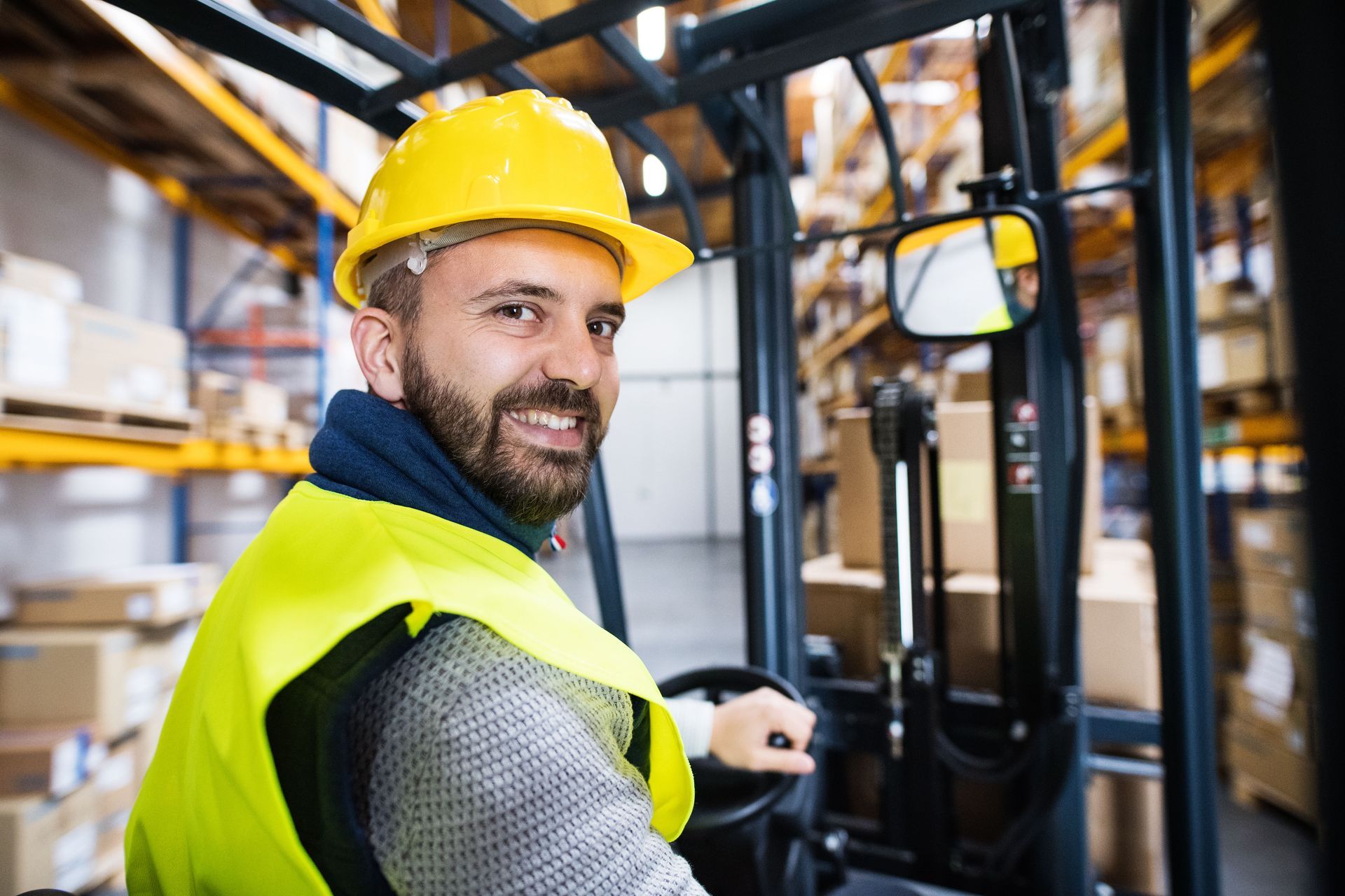 A man wearing a hard hat and safety vest is driving a forklift in a warehouse.