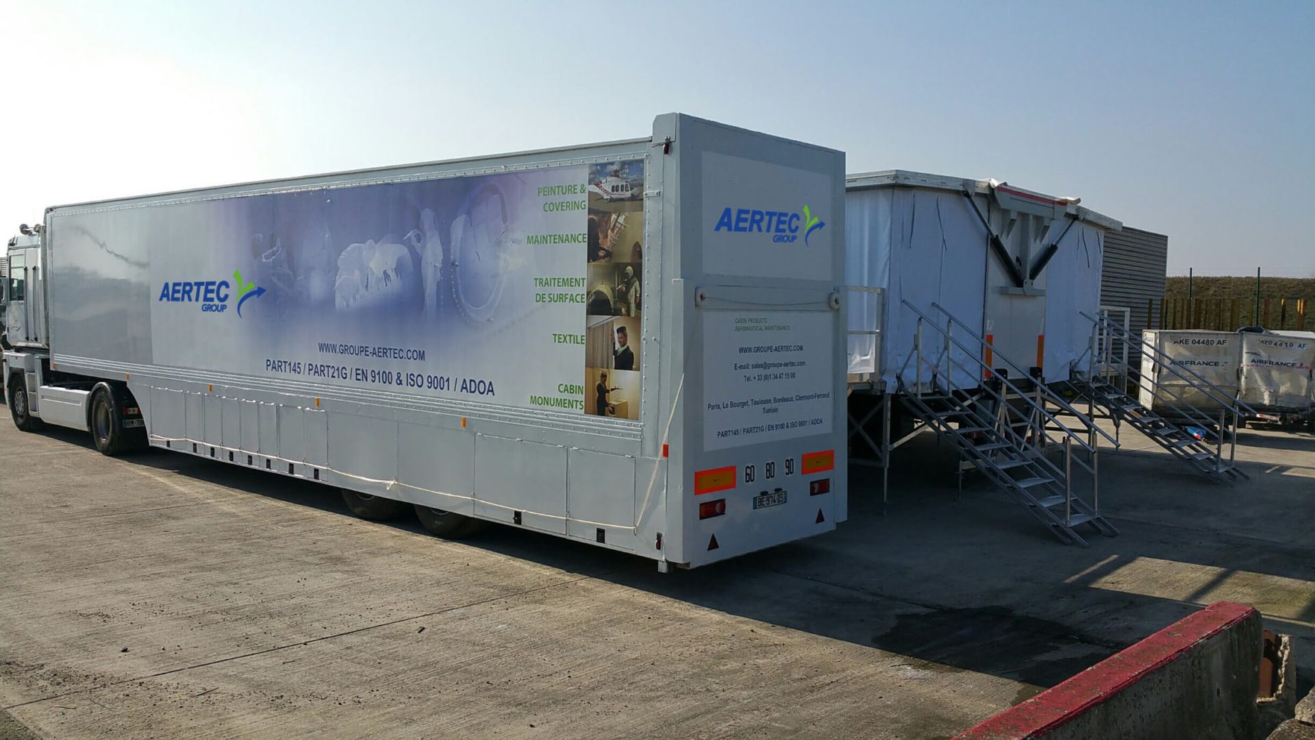 Semi-remorque blanche portant le logo « AERTEC ». Une autre structure se trouve à droite, sur une surface en béton.