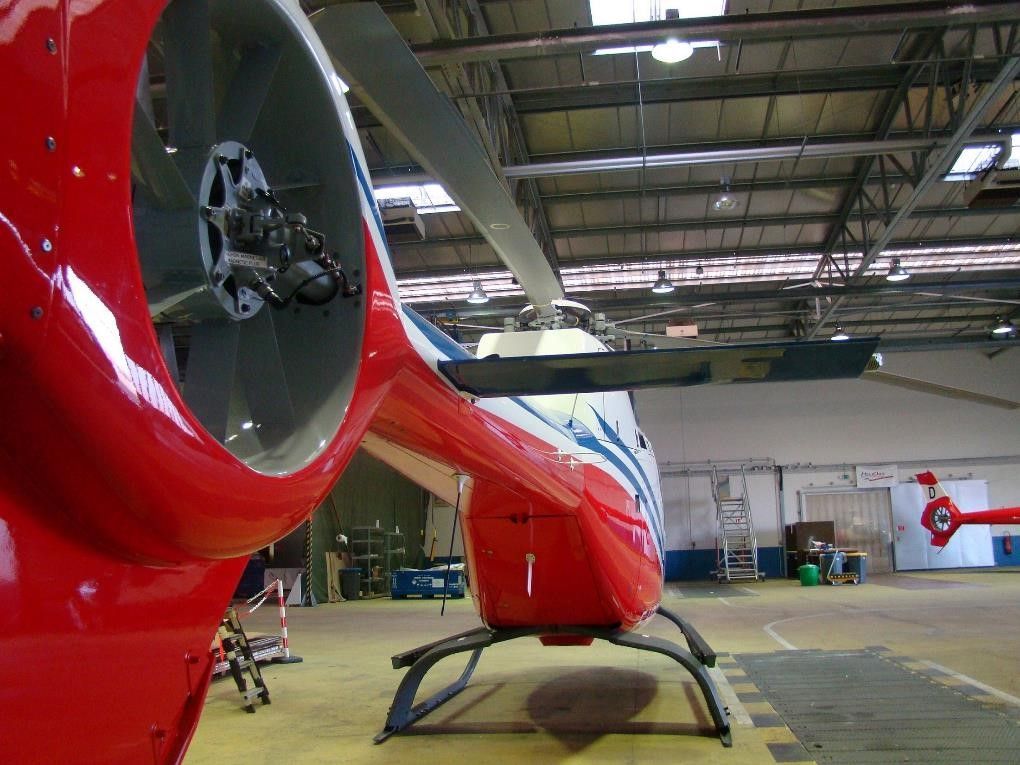 Hélicoptère rouge et blanc dans un hangar, vu de l'arrière, rotor de queue visible.