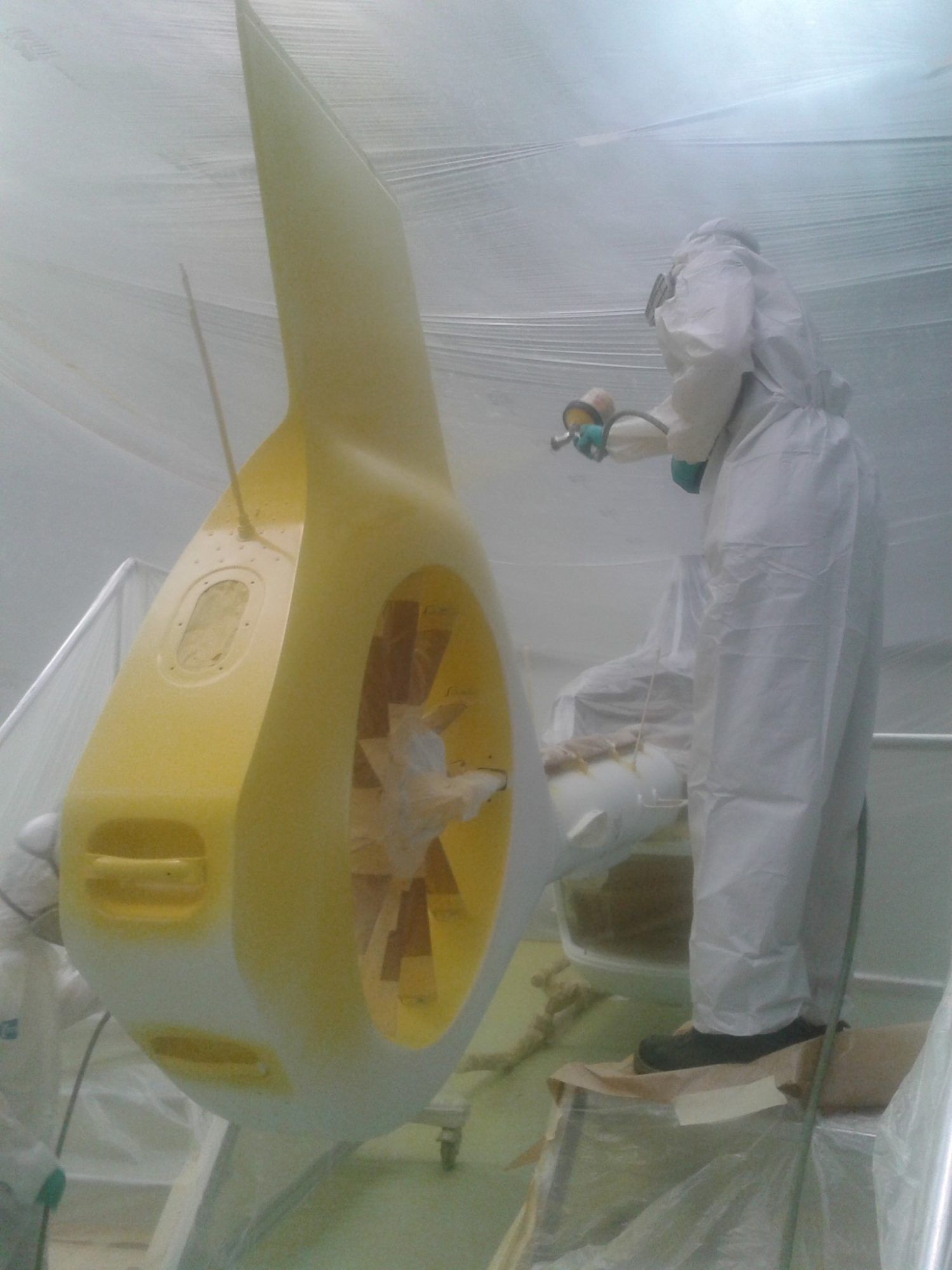 Une personne en combinaison de protection peint à la bombe un grand objet jaune dans une pièce blanche.