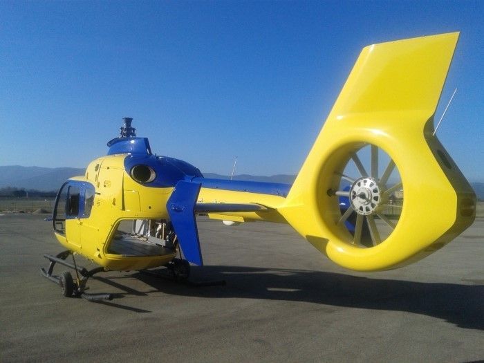 Hélicoptère jaune et bleu au sol, protection du rotor de queue incluse, journée ensoleillée.
