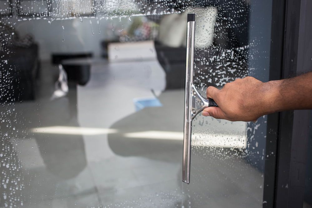 A Person Cleaning a Glass Door with A Squeegee, Covered in Soapy Water — EDU-BABA Pressure Cleaning in Weston Creek, ACT
