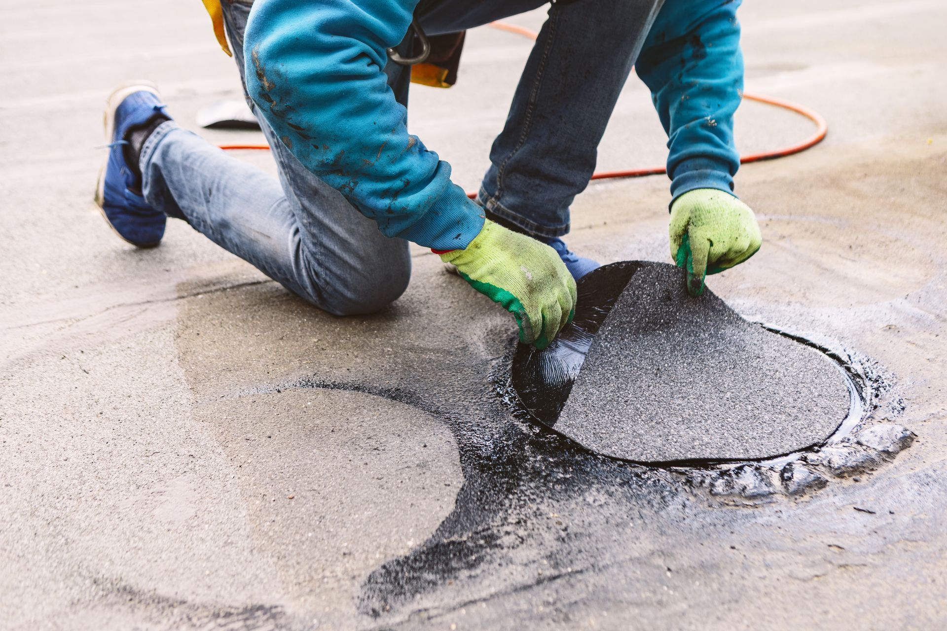 Person in blue jacket and jeans kneels, applying patch to a roof. Wearing gloves.
