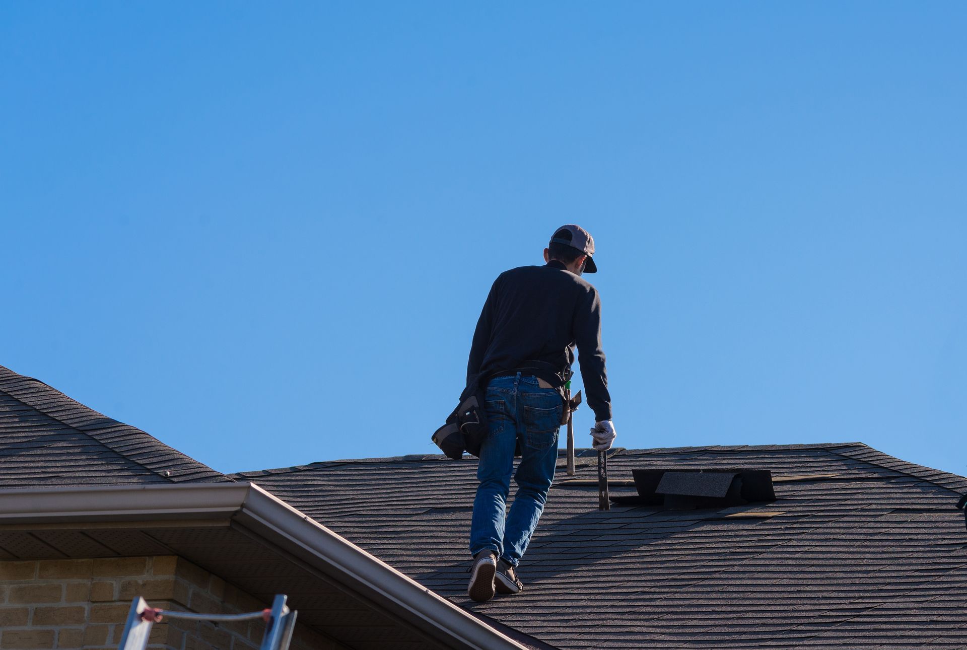 Roofer on a dark shingled roof under a clear blue sky, working with tools.