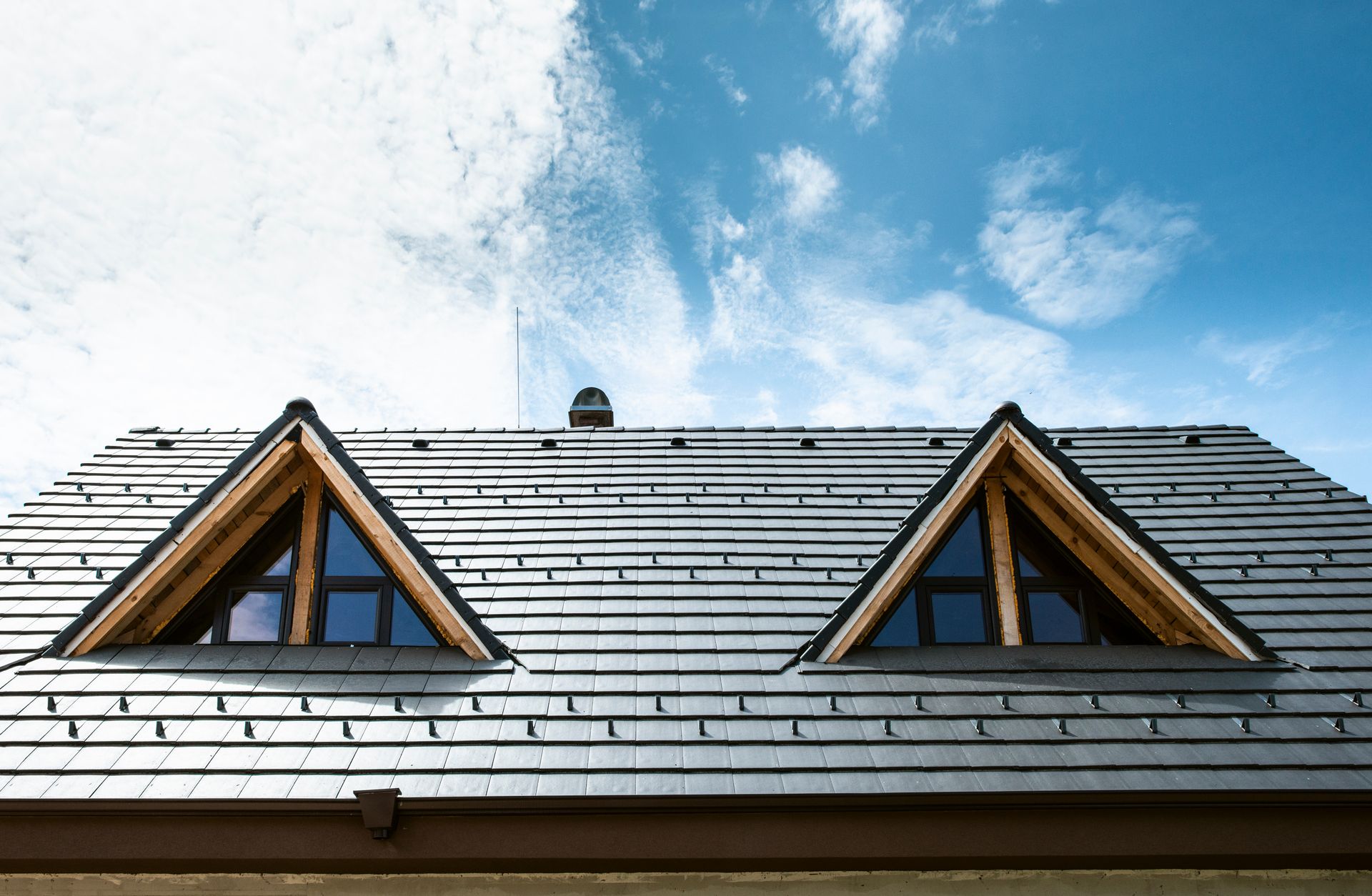 A slate roof with two triangular windows, under a partly cloudy sky.