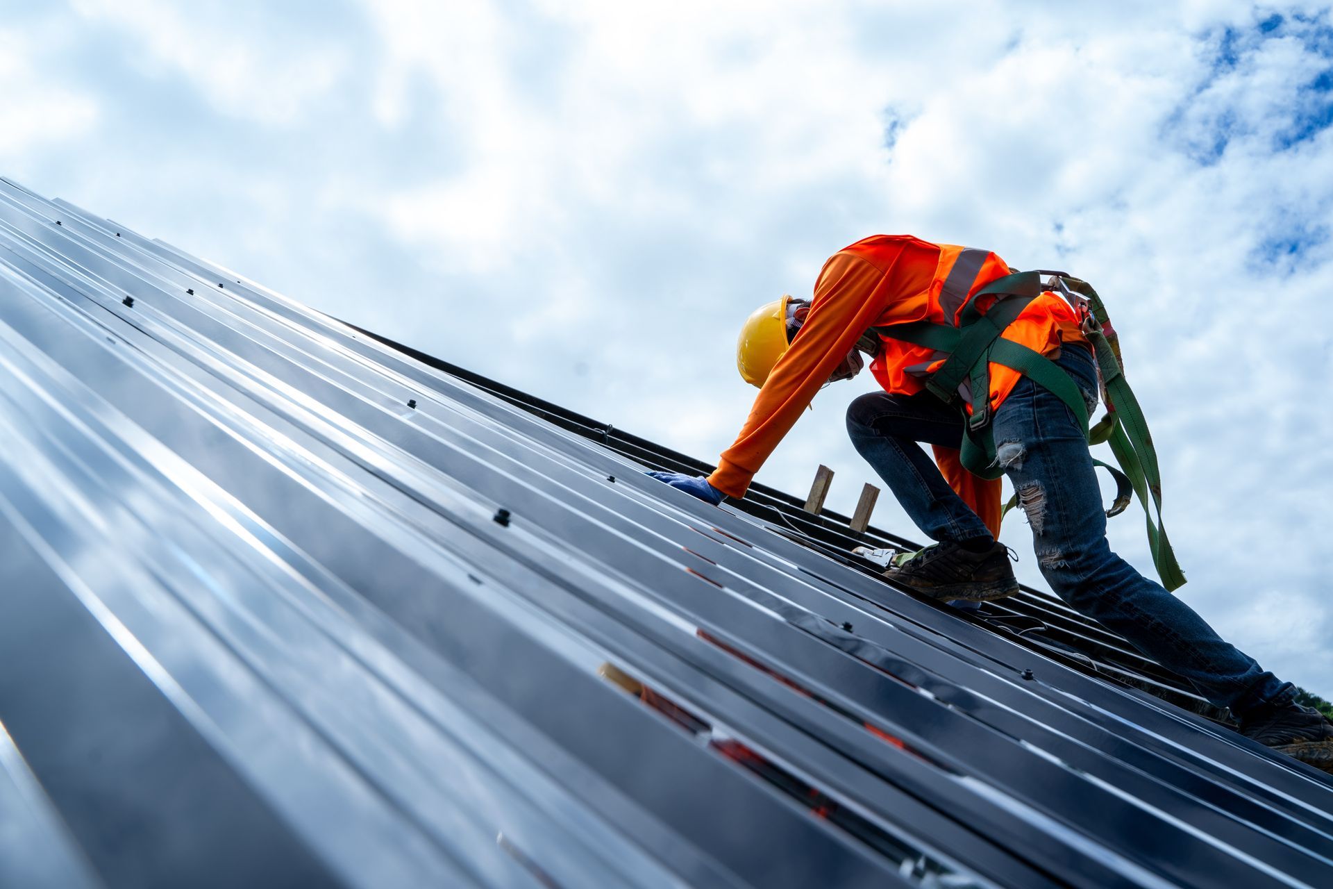 Roofer in orange safety vest and helmet installs metal panels on a roof, secured by a harness. Cloudy sky.