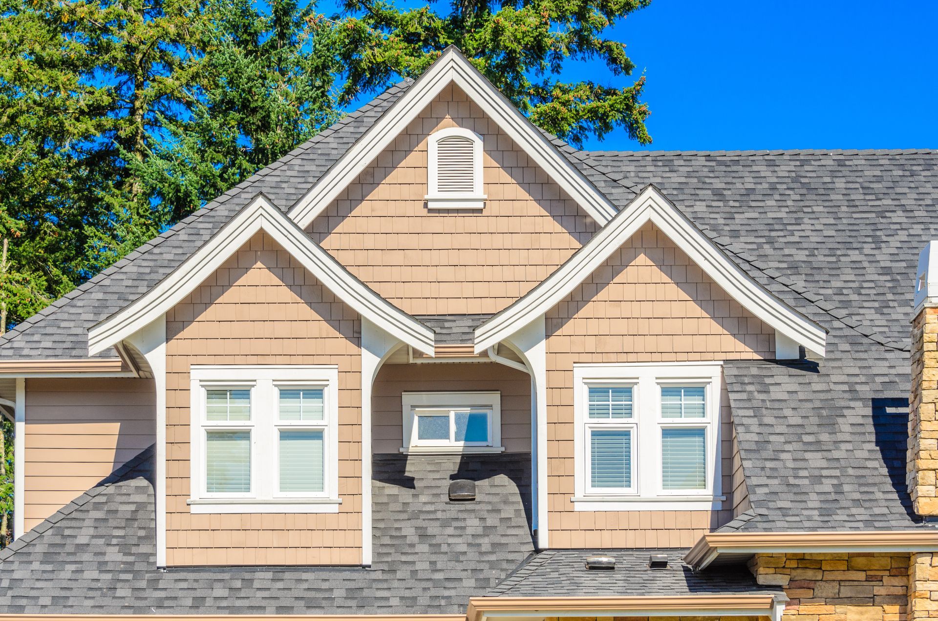 Tan and grey house exterior with gabled roof, windows, and light siding against a blue sky.