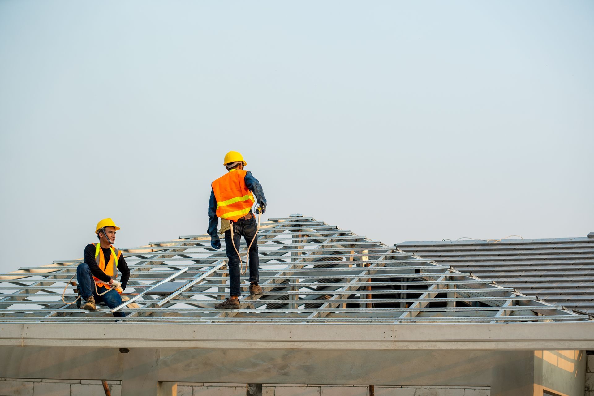 Two construction workers wearing safety vests and hard hats on a roof frame.