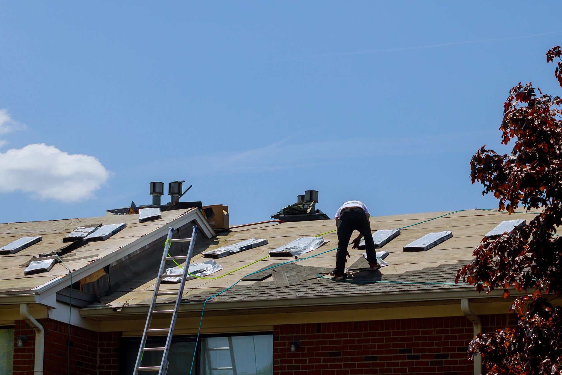 Person working on a roof, installing new shingles under a blue sky. A ladder stands nearby.