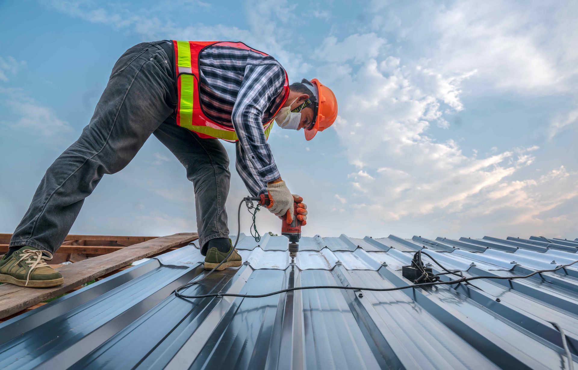 Roofer on a metal roof, wearing safety gear, drilling with a power tool against a cloudy sky.