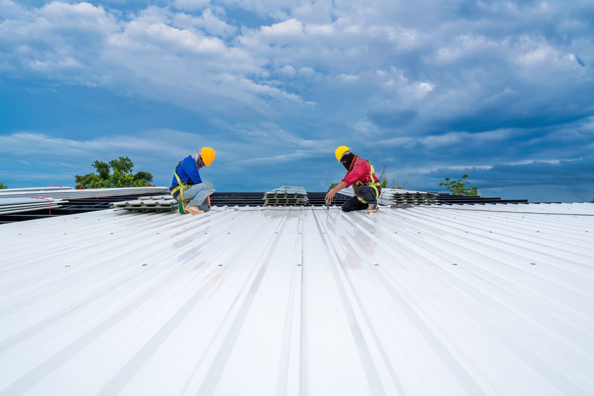 Two workers on a white metal roof, wearing harnesses, under a cloudy blue sky.