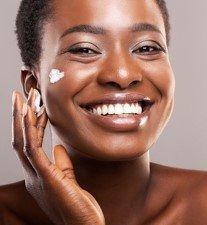 Smiling person applying white cream to cheek against a neutral background