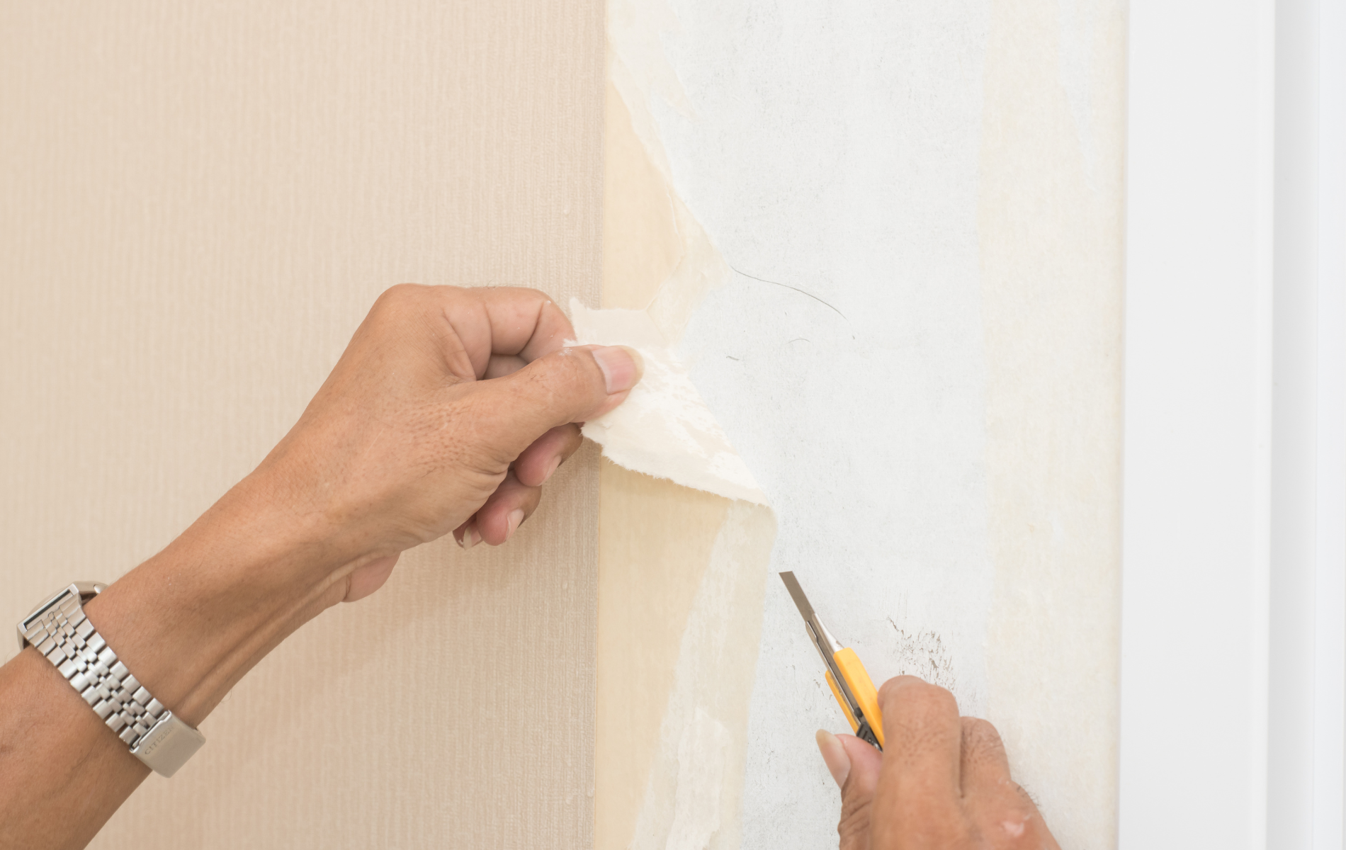 Person peeling wallpaper with a knife on a beige wall, next to a white door frame.