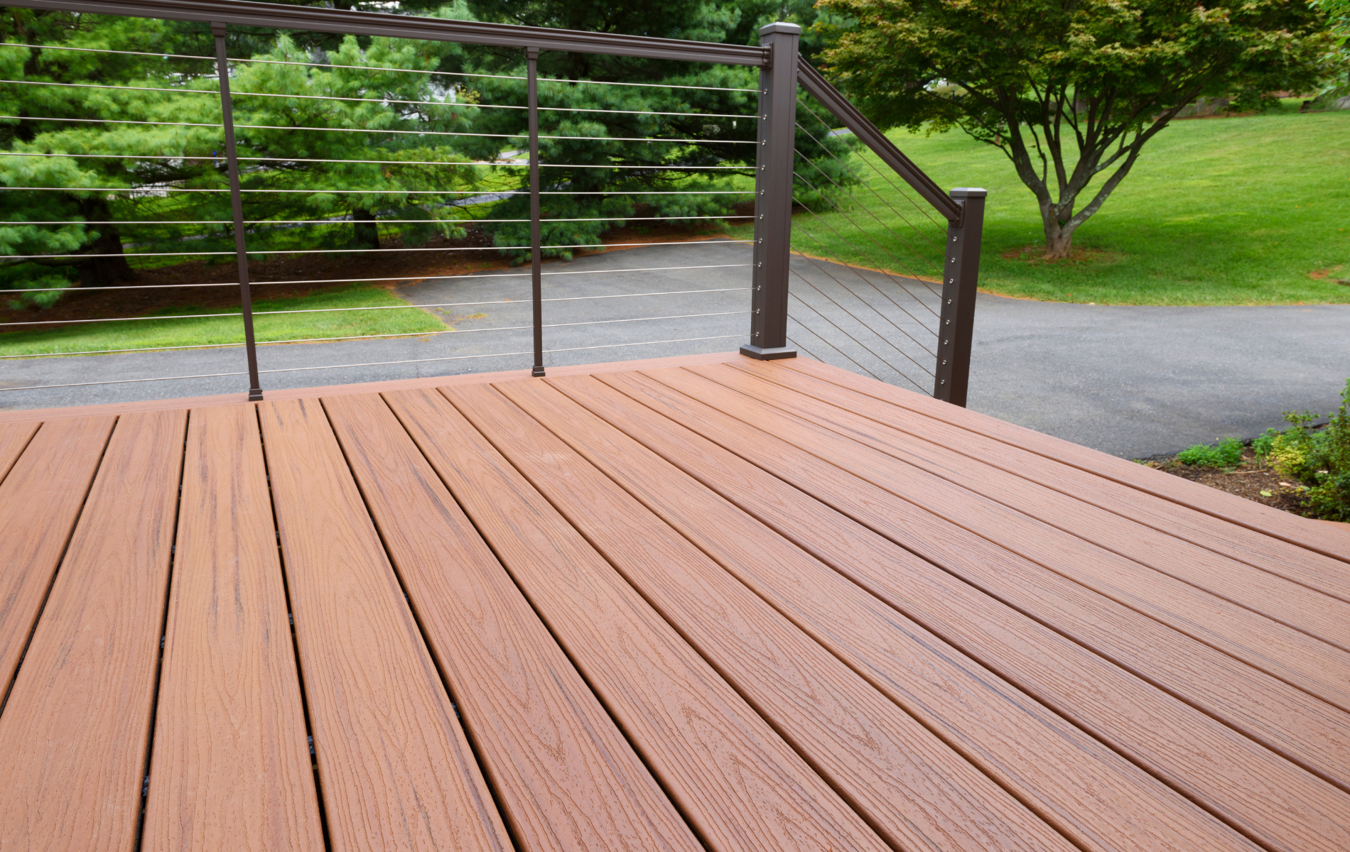 Wooden deck with cable railing, overlooking a paved path and green yard.