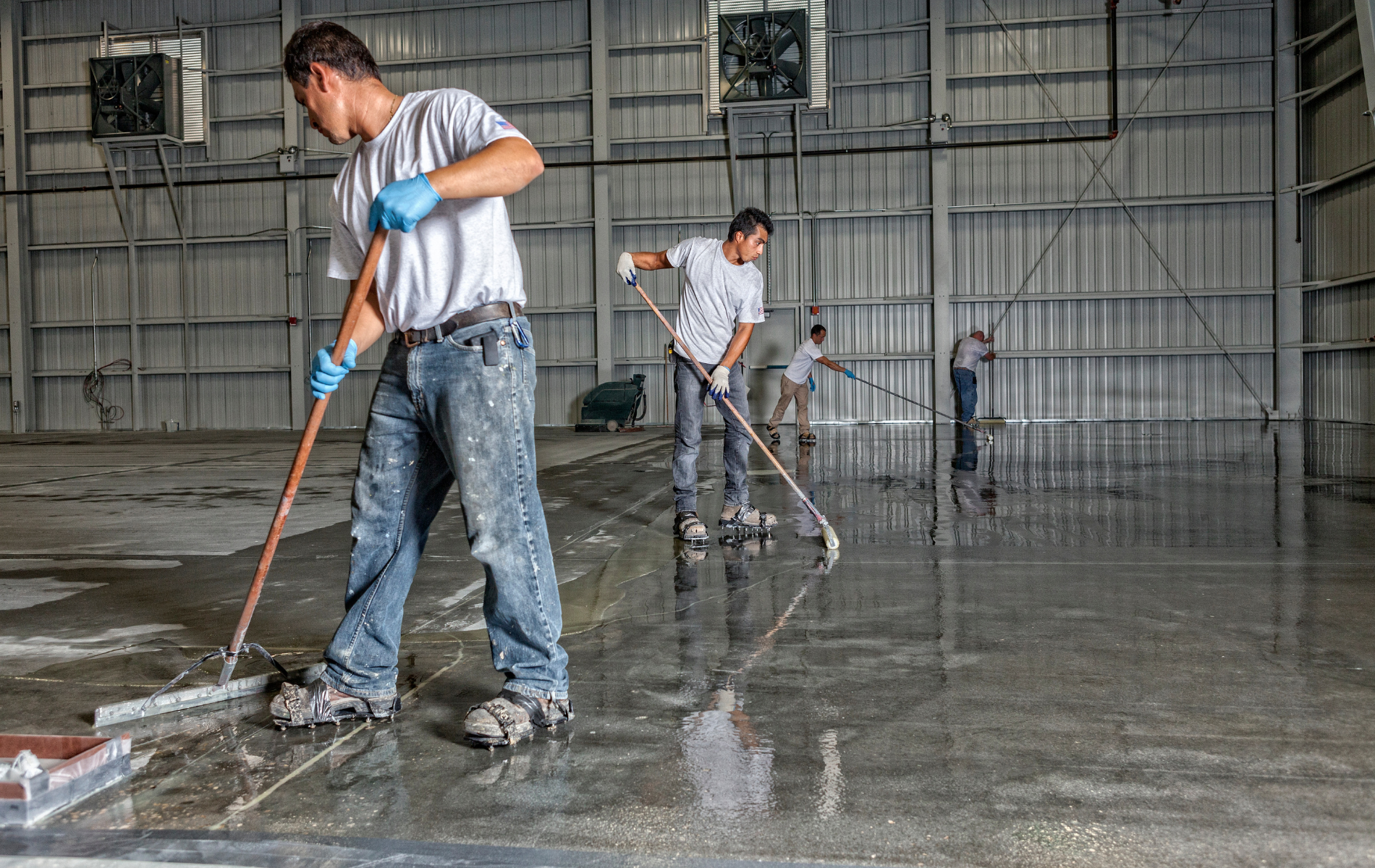 Men in white shirts and jeans applying sealant to a concrete floor in a large industrial building.