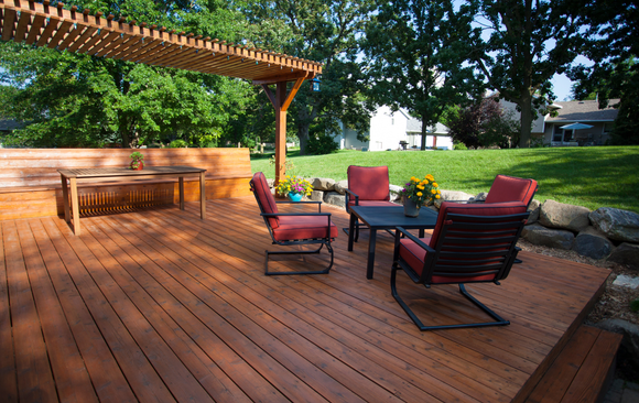 Wooden deck with red chairs, a table, and a pergola. Overlooks a lawn with trees.