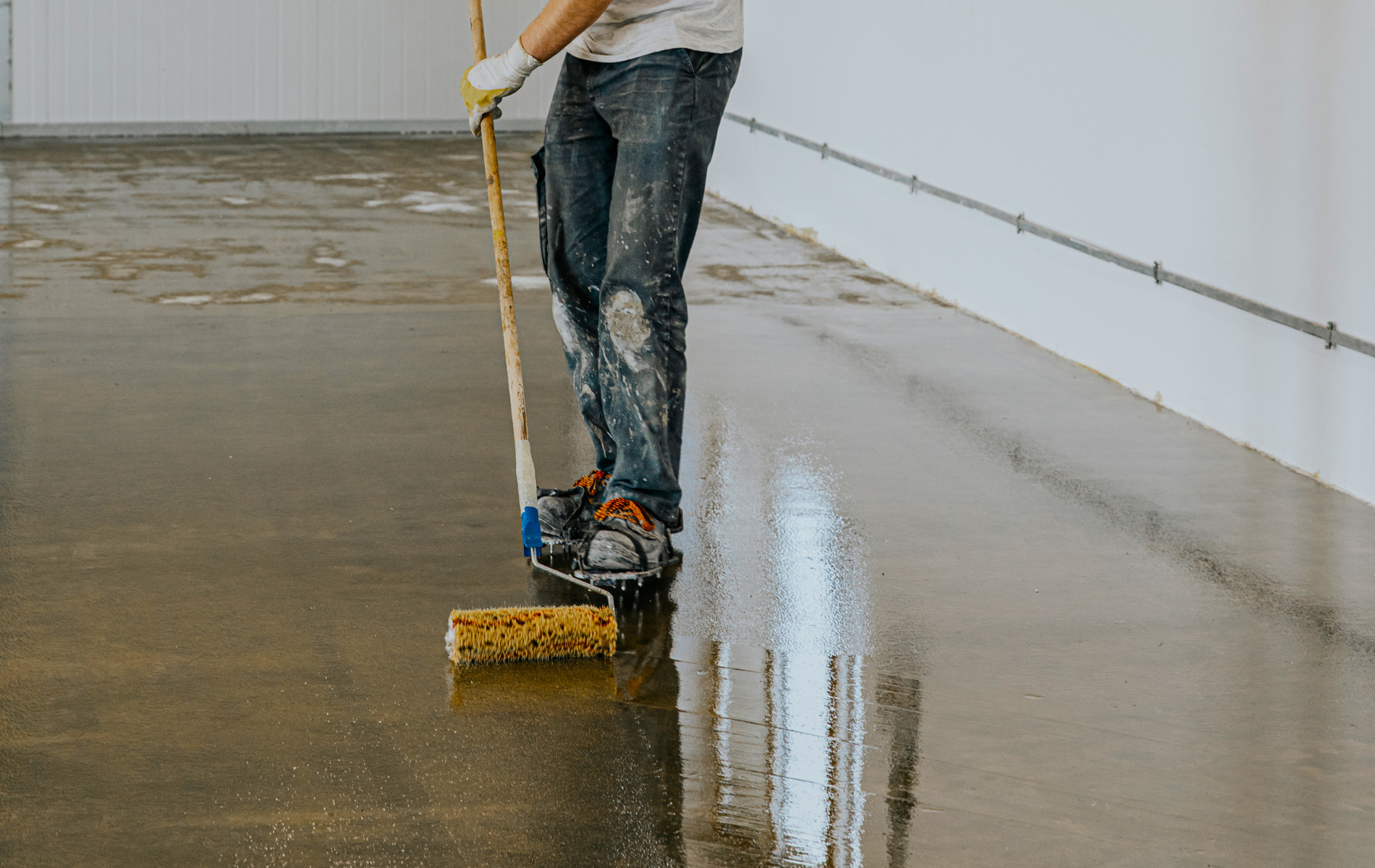 Person applying a sealant to a concrete floor with a roller in a white-walled room.