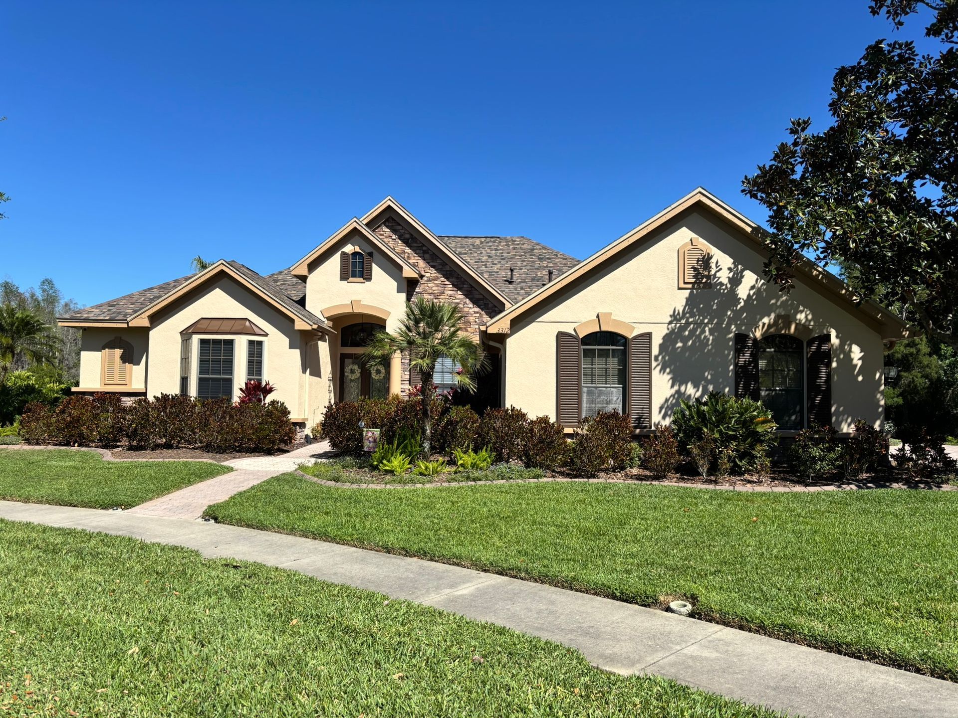 A beige and brown house with a brick facade, green grass, and a sidewalk under a blue sky.