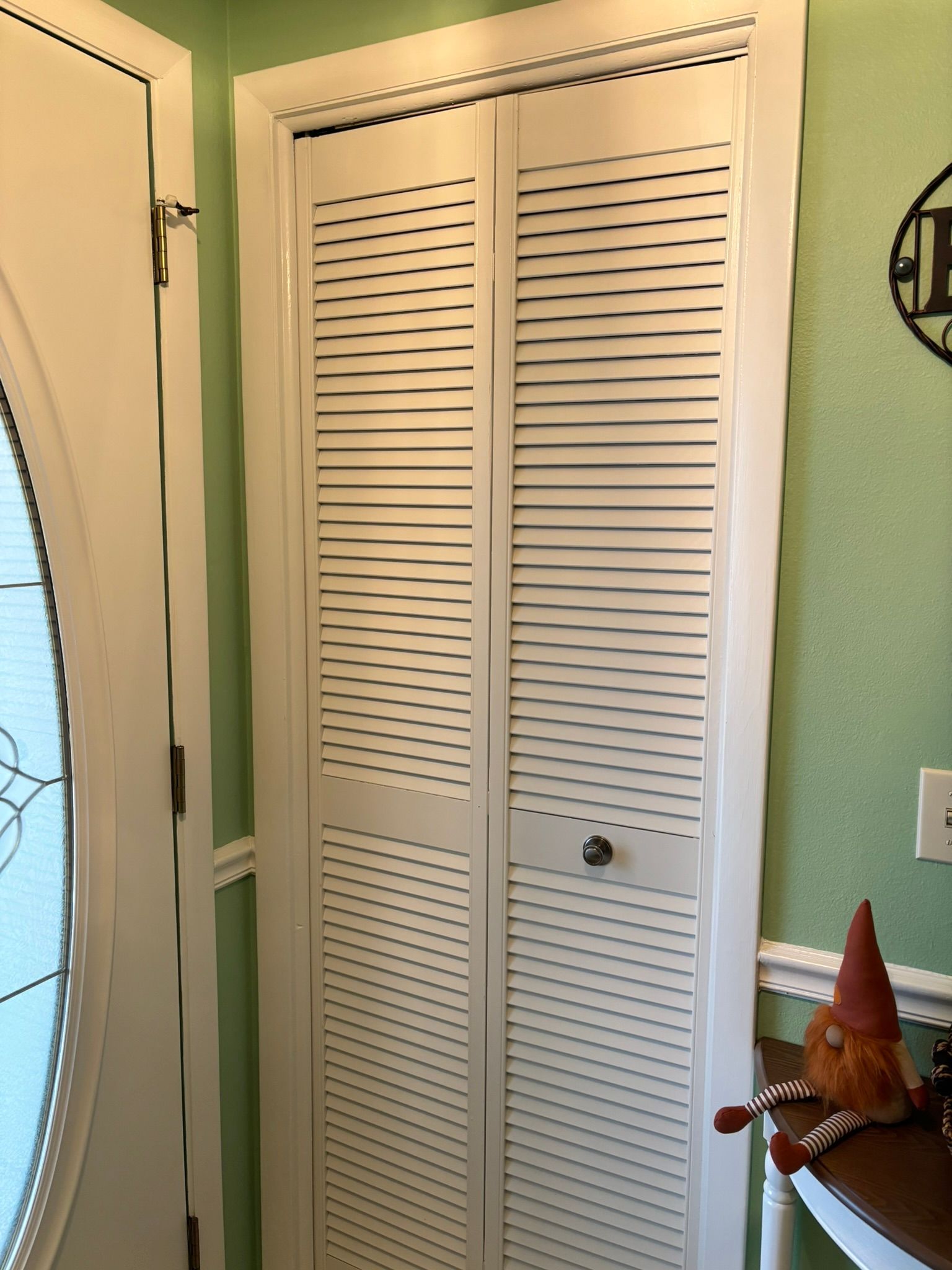 White louvered bi-fold closet door in a corner, next to a door with oval glass. Green wall.