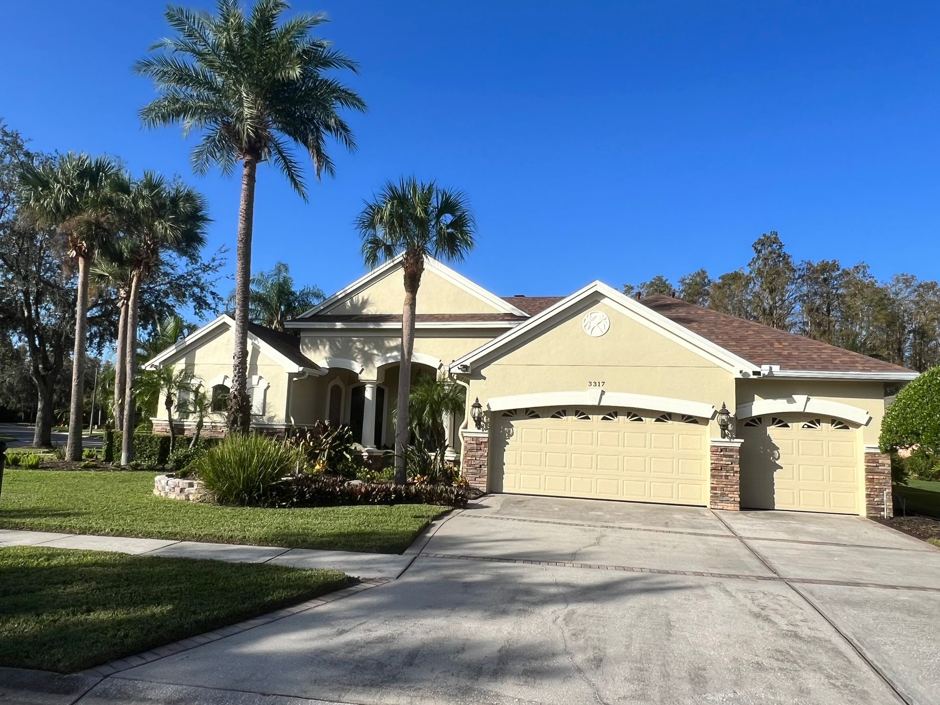 Beige house with brown roof, three-car garage, and palm trees in front on a sunny day.