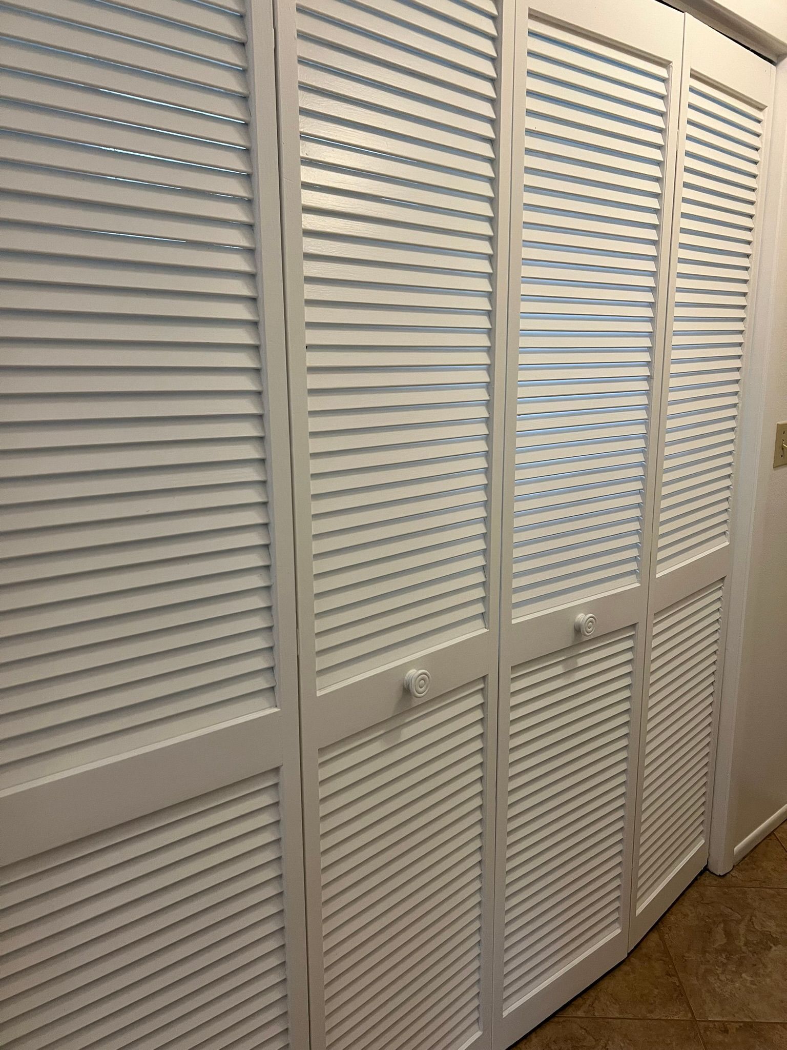 White louvered closet doors, closed. Handles visible. Brown flooring, white wall.