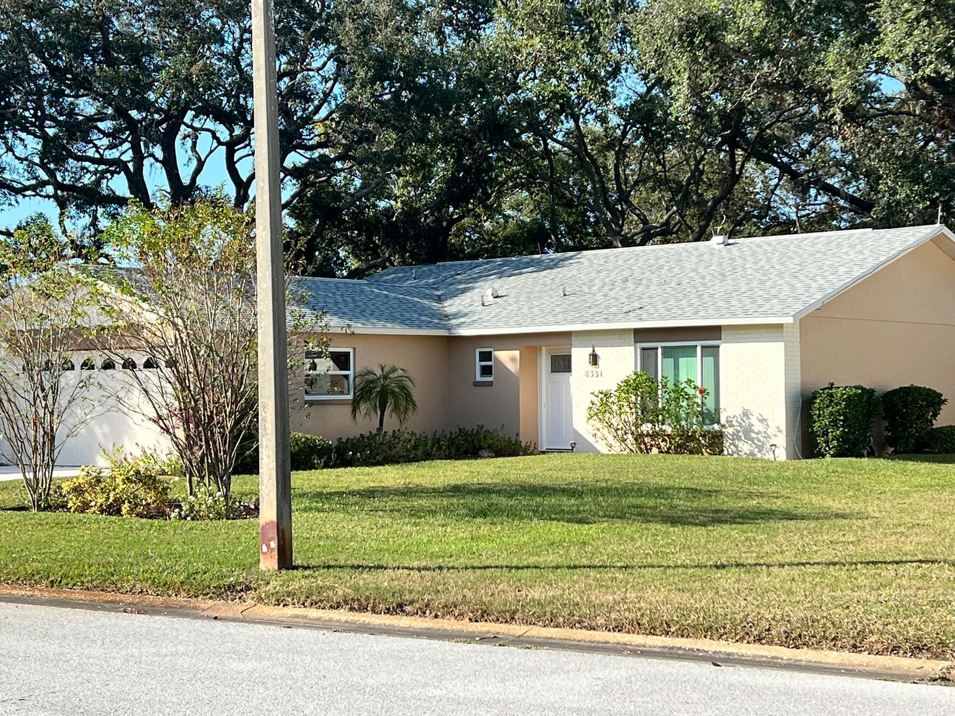 Beige ranch-style house with a light gray roof, small front yard, and tall trees in the background.