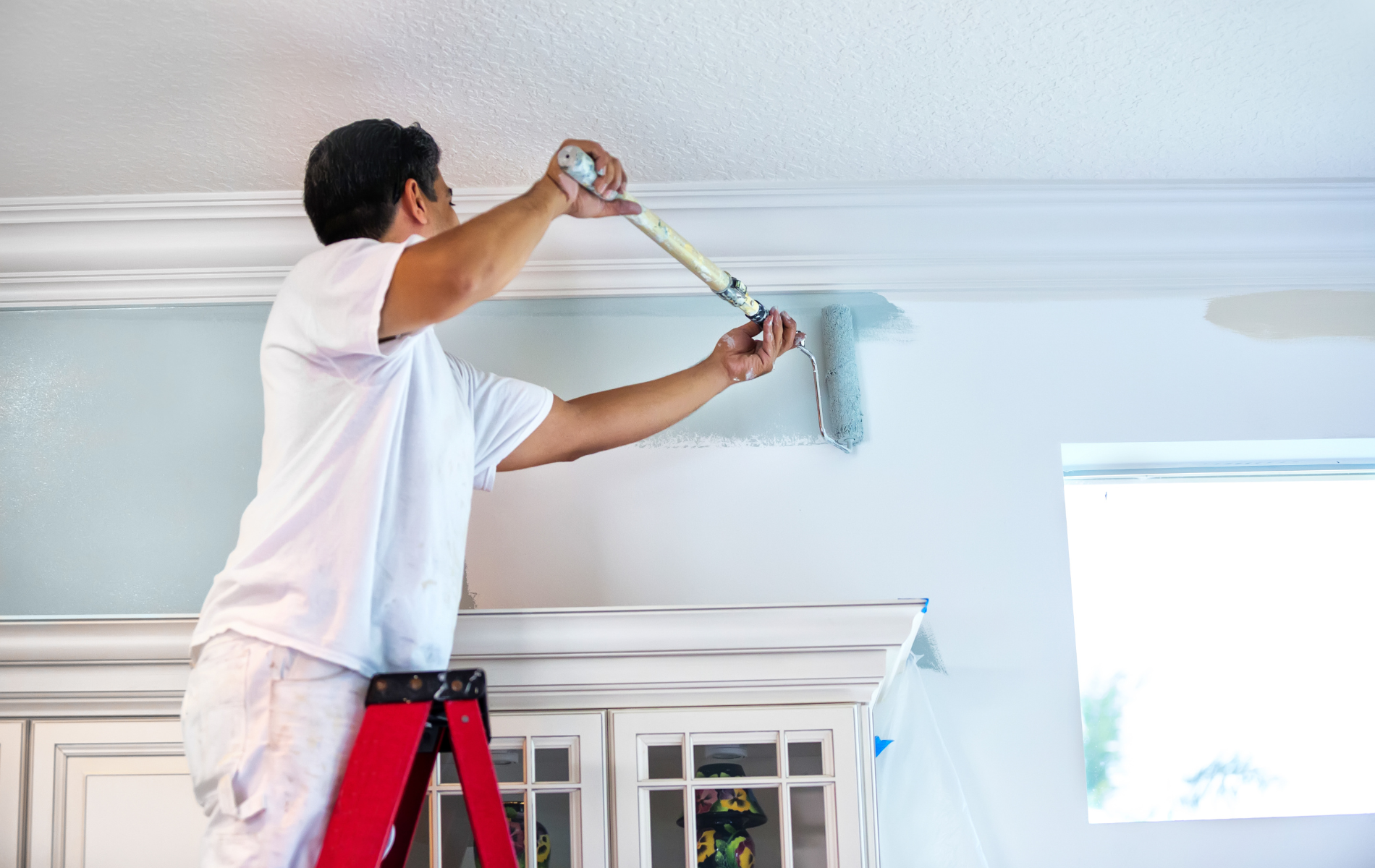 Man on a ladder painting a wall with a paint roller, near a decorative trim.