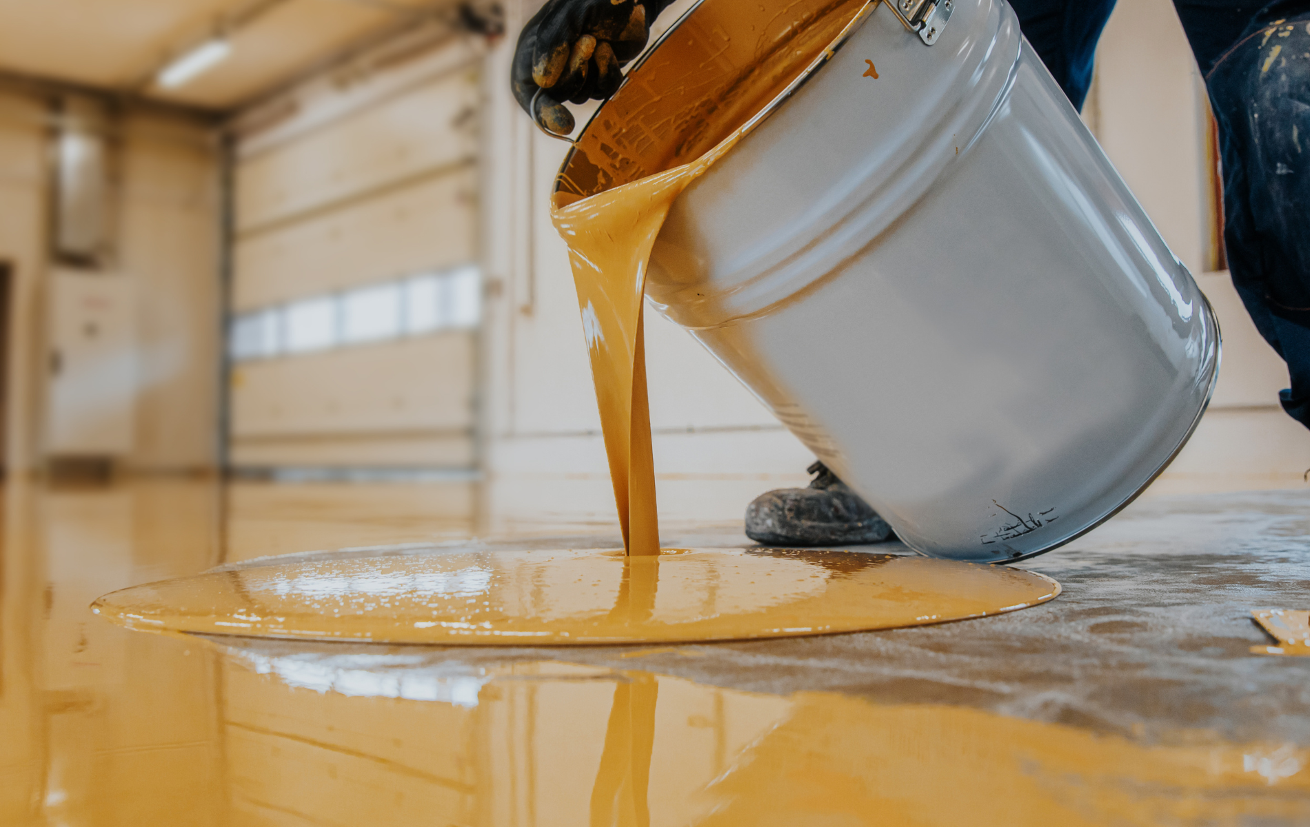 Yellow liquid epoxy being poured from a bucket onto a concrete floor in a warehouse.