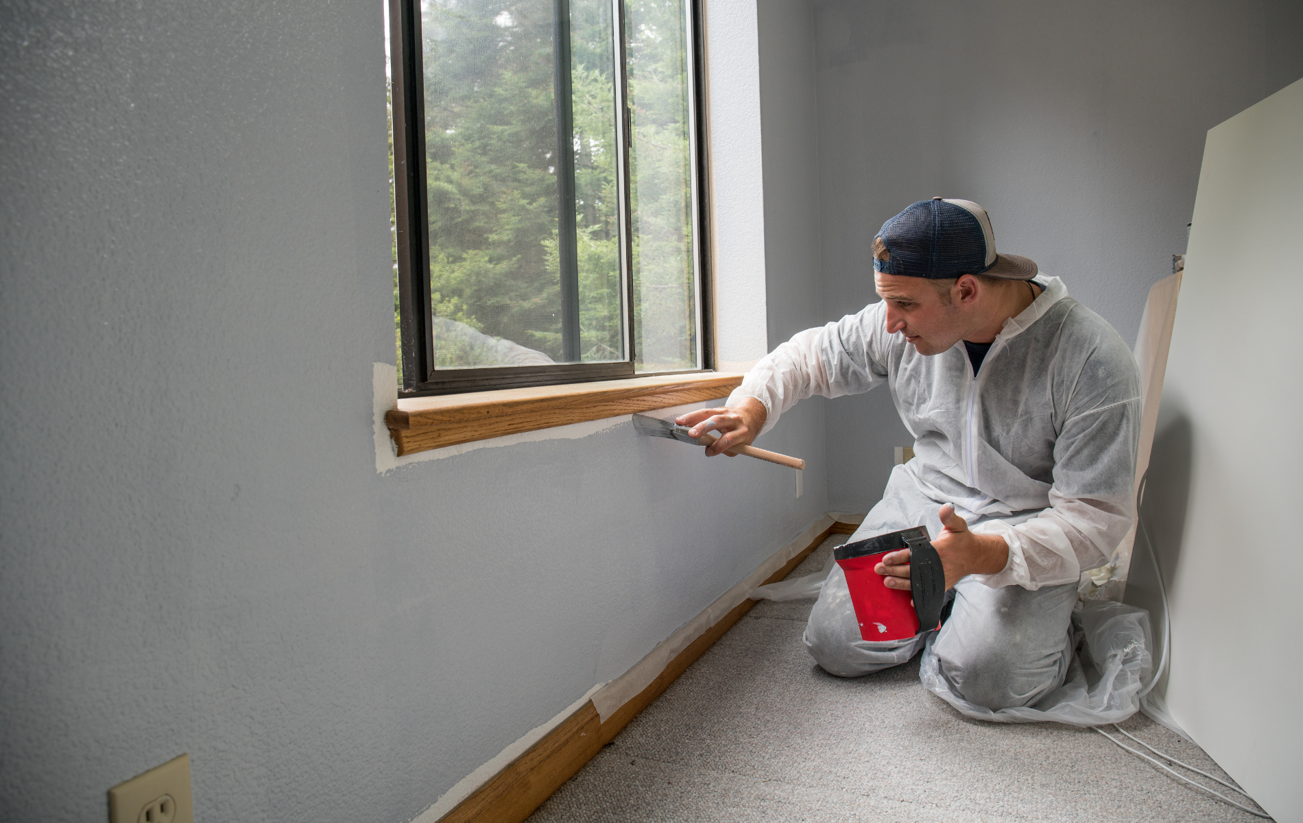 Man in protective suit kneeling, applying putty around a window frame. Gray walls, brown wood trim.