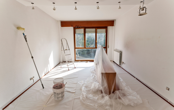 Interior of room being painted, white walls and floor covered, ladder near window.