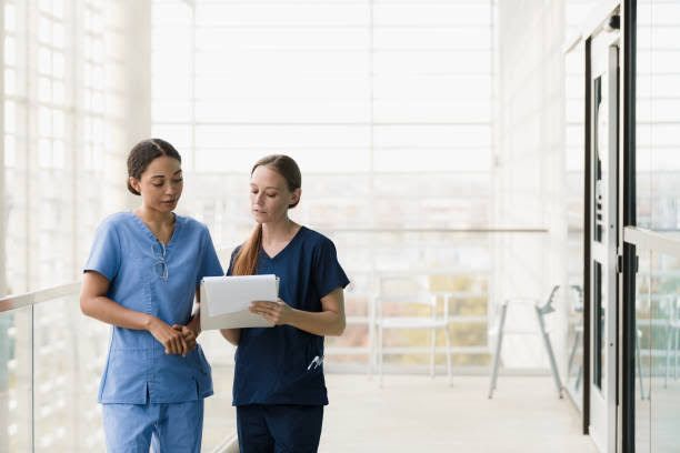 Two nurses are looking at a tablet in a hospital hallway.