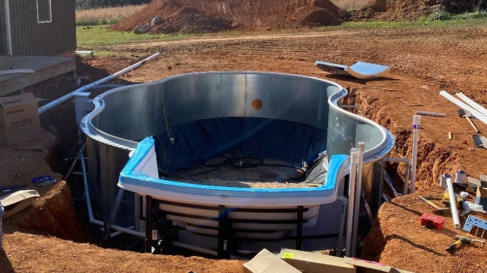 Pool construction site: partially installed above-ground pool in an excavated area, surrounded by dirt and construction materials.