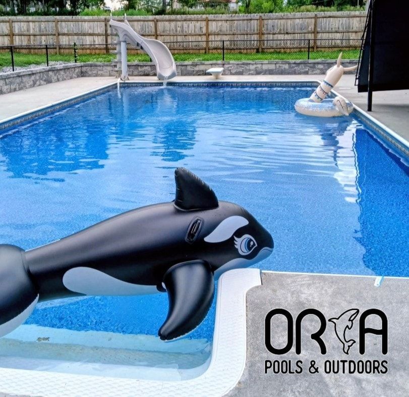 A backyard swimming pool with an inflatable orca in the foreground, a slide in the distance, and a brand logo in the corner.
