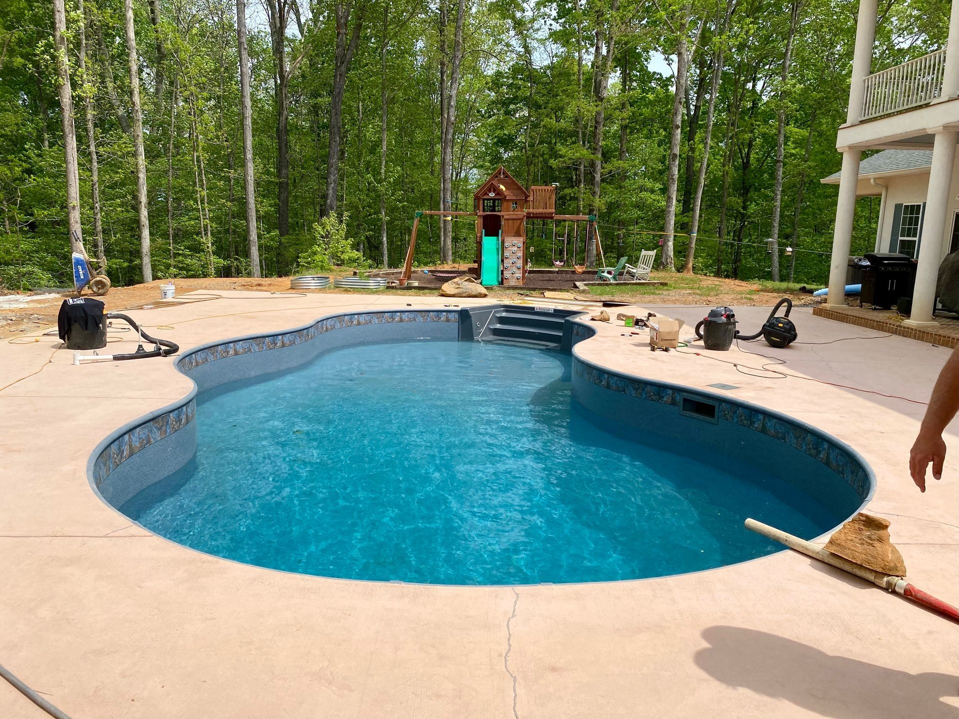 A partially filled swimming pool with concrete deck, surrounded by trees. A playground sits in the background.