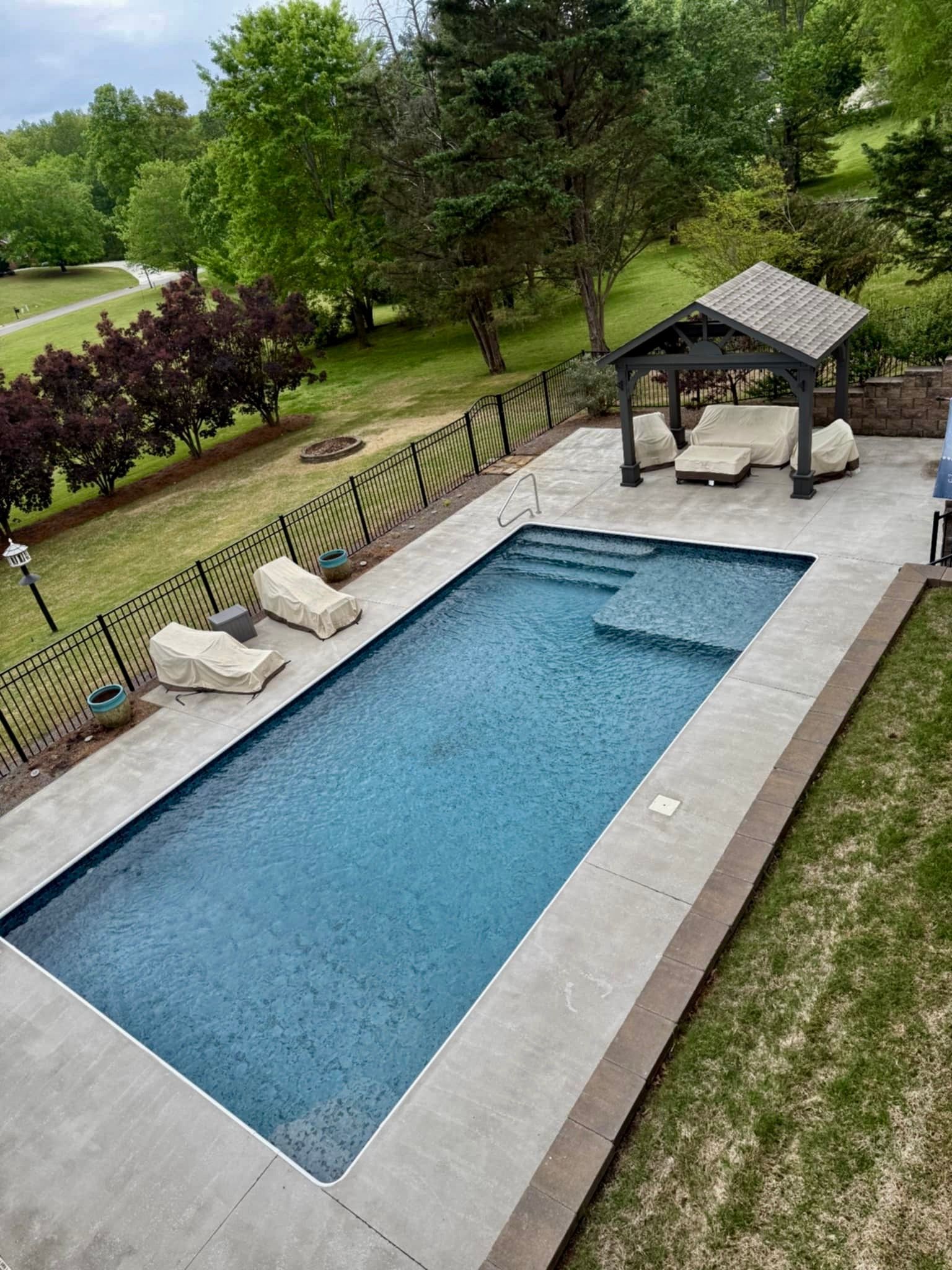 A rectangular pool with a gazebo, surrounded by concrete, grass, and a black fence.