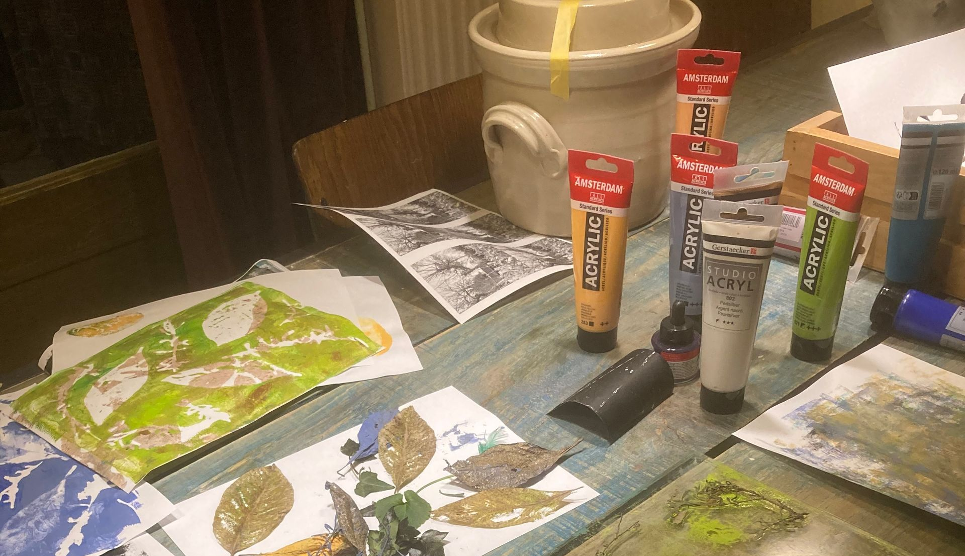 A table with art supplies: paint tubes, prints of leaves, and a ceramic pot.