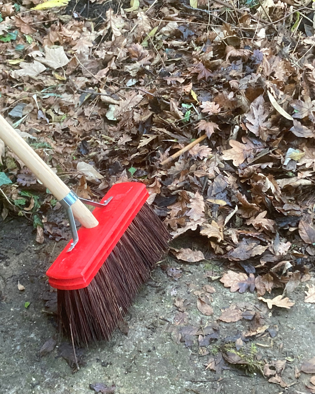 Red broom sweeping dried leaves on a concrete surface.