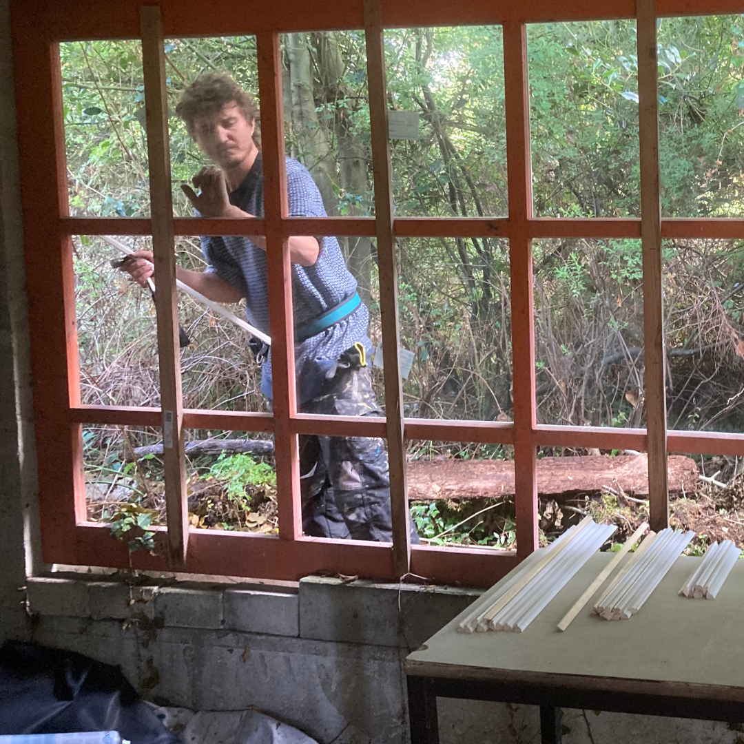 Een man staat buiten bij het raam, met gereedschap in zijn hand, en werkt aan een kozijn. Binnen liggen witte stokken op een tafel. Op de achtergrond is een bos te zien.