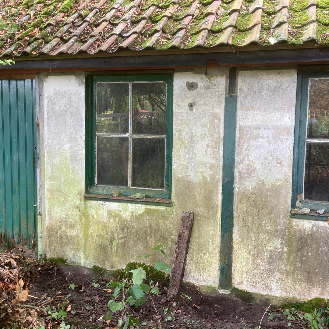 Oud, verweerd gebouw met groene kozijnen, een met mos begroeid dak en een vervallen buitenkant.